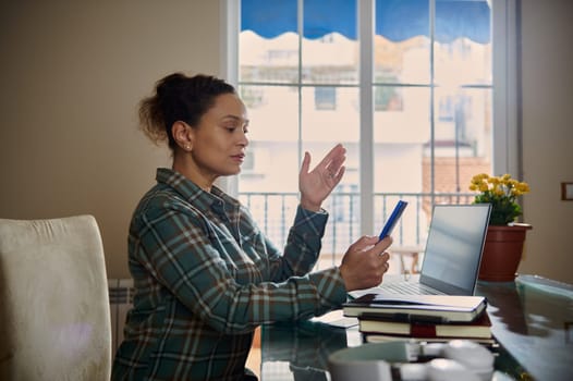 A woman engaged in remote work, utilizing her smartphone and laptop to stay connected and productive in a home office setting that emphasizes modern technology and flexibility.