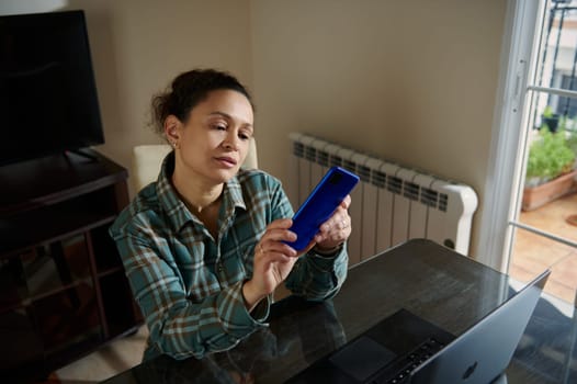 A woman sits at her home office table using a smartphone, with a laptop nearby. Bright and cozy indoor setting emphasizes focus and connection, bringing a sense of productivity and comfort.