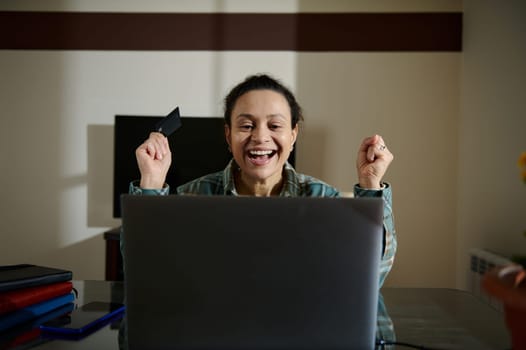 A joyful woman celebrates from a successful online transaction using her credit card and laptop, sitting at a desk. This image reflects happiness, success, and the convenience of online shopping.