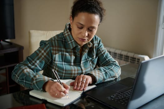 A woman focuses on her tasks, using her laptop and writing notes, seated in a comfortable home workspace. The image reflects a dedicated and productive atmosphere.