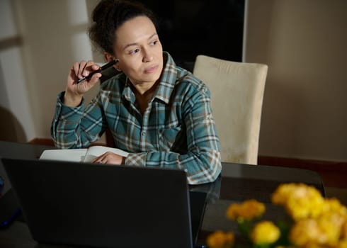 A woman sits at a desk with a laptop and notebook, focusing on her work in a cozy home setting. Thoughtful and engaged, she appears to be planning or writing ideas.