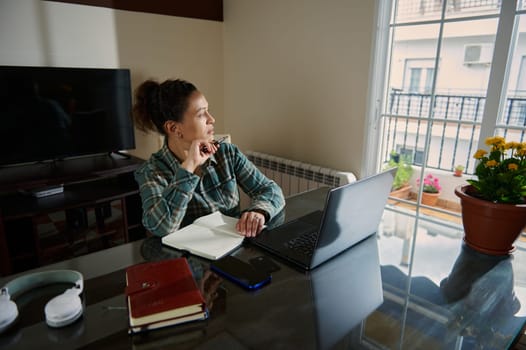 Woman working from home on a laptop near a window, looking introspective, balancing productivity and creativity in a relaxed atmosphere with a touch of nature from indoor plants.