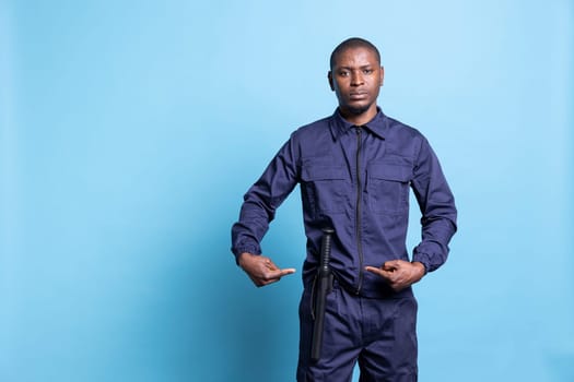 Armed security officer presenting his baton weapon for own safety in studio, pointing at his police bat to ensure order and protection. Male trained personnel in uniform against blue background.