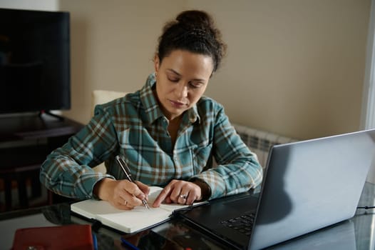 A focused woman writes in a notebook while working on a laptop in her home office. The scene depicts concentration, organization, and productivity in a comforting and cozy environment.