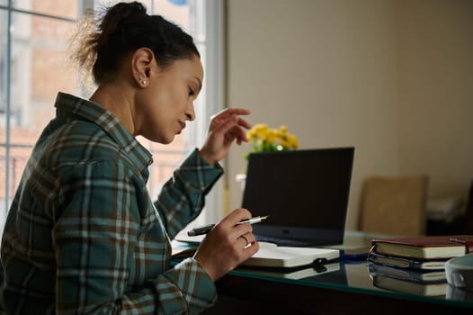 A woman intently focused while writing notes in a notebook, seated at a home office desk with a laptop and other tools for work, indicating productivity and dedication.
