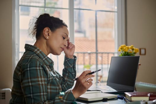 A woman focuses intently while working at home, seated by a window with her laptop, notebook, and pen. The environment suggests concentration, remote work, and a productive home office setting.