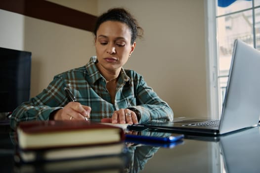 A woman working at a desk, writing notes while using a laptop computer. She is in a home office environment, multitasking and focusing on productivity.