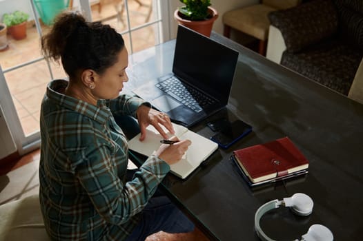 A focused woman takes notes in a journal while working from her home office. Her workspace includes a laptop, books, and headphones, reflecting organization and dedication to her remote work.