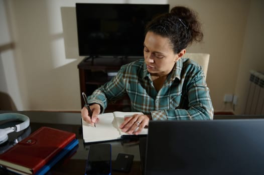 A determined woman diligently writes notes while multitasking on her laptops, showcasing productivity and focus from the comfort of her home office.