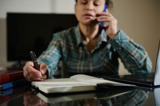A woman multitasking in a home office environment, handling conversations over the phone while making written notes in a planner. A scene of productivity and focus, representing professional multitasking.