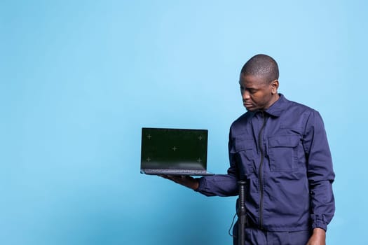Male security agent pointing at his laptop screen with mockup, presenting a greenscreen while he poses in studio. Young skilled guard standing against blue background, patrol service.