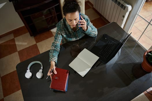 Overhead view of a focused woman multitasking at a desk with a laptop, notebook, headphones, and smartphone. The setting suggests productivity and remote work in a home office environment.