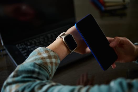 A person wearing a smartwatch interacts with a smartphone in a modern desk workspace. The technology-focused setting highlights contemporary communication and digital smart devices.