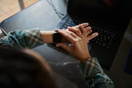 Midday close-up of hands checking smartwatch while working on a laptop in a workspace, highlighting technology integration in daily productivity. Captures modern work-from-home or remote work lifestyle.