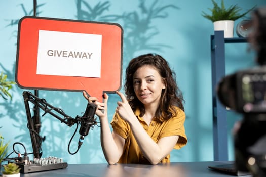 Joyous influencer pointing to empty copy space sign advertising giveaway for fans. Woman offering prizes during social media contest, filming with professional camera, holding cutout