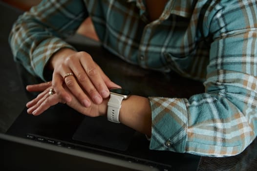 A close-up of hands resting on a smartwatch over a laptop. The individual wears a flannel shirt, emphasizing a relaxed home office setting and symbolizing modern technology usage.