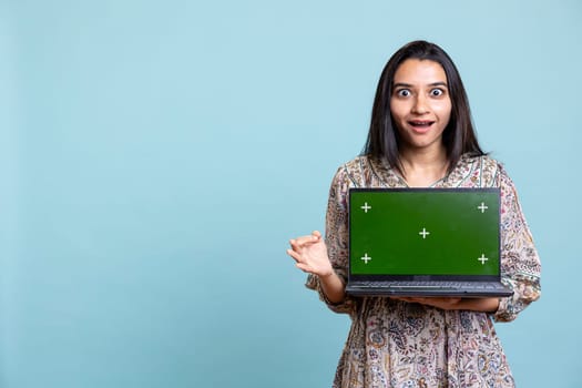 Female influencer presenting green screen on a gadget in studio, standing against studio background. Young enthusiastic person using copy space display for brands advertisement.