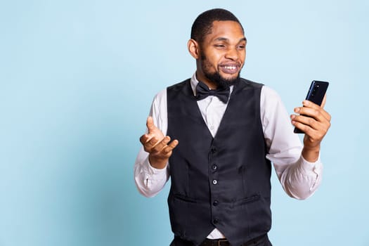 Doorman with suit and tie answering online videocall on phone, discussing with clients or guests against blue background. Bellboy staff on videoconference connection in studio.