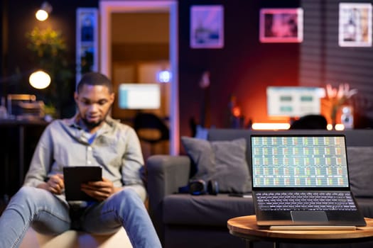 Black man day trading from home on laptop following stock market charts and financial graphs. Remote work setup shows an investor tracking prices, trends and data for online investment decisions.