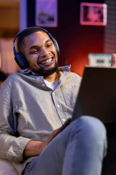 Confident male person attends a webinar lesson online on a video call, paying attention and engaging in discussion remotely. Young black guy meeting with attendees on laptop, relaxing on sofa.
