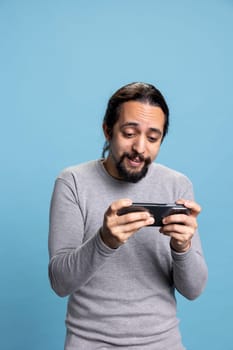 Casual modern guy playing video games tournaments on mobile phone, participating in an electronic competition of a shooter game against blue background. Young person with long hair and beard.