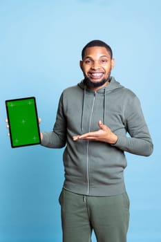 Happy influencer holding mockup chroma key display on tablet, doing recommendation and talking with the audience. Cheerful guy pointing at copy space screen against blue background.