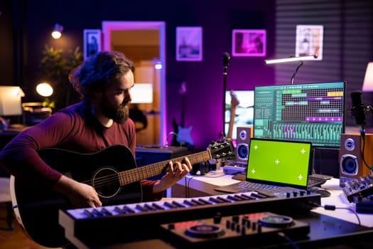 Artist songwriter playing guitar by watching online musical lessons on laptop with greenscreen, learning to play acoustic instrument in home studio. Musician singing to record tunes.