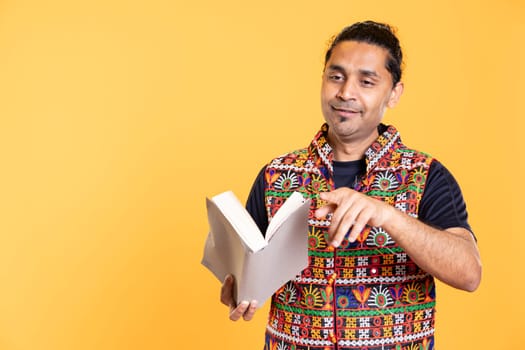 Jolly indian teacher reading book for pupils during class, pointing finger gesture, studio background. Happy man holding novel, telling story to kindergarten kids, asking them questions