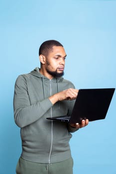 Carefree happy african american guy working on his laptop in studio, wearing a grey hoodie and composing emails to solve his tasks and paperwork. Young adult multitasking, browses websites.