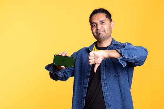 Confident guy doing the thumbs down sign next to mockup on mobile phone, creating an advertisement for a promotion against studio background. Indian man does hand gesture.