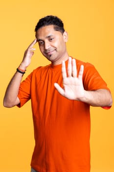 Indian friendly male person with a man bun raising his hands and waving, greeting his friends against orange background. Young adult in a casual clothes, joyous satisfied guy.