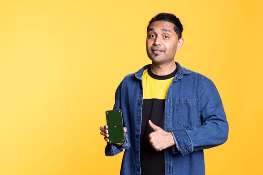 Indian male model presenting mobile phone with green screen in studio, posing against studio background. Young person dressed in casual attire and shows thumbs up sign, positive mood.