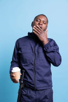 African american security guard feels sleepy after a night shift, serving a cup of caffeine beverage to deal with exhaustion. Male officer working as bodyguard, yawning against blue background.
