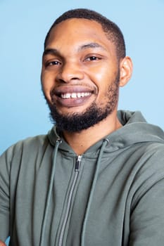 Portrait of african american person in casual grey hoodie smiling, posing in studio and feeling happy. Young carefree jolly adult being positive against blue background, show emotions.