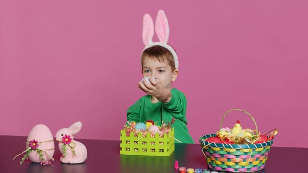Excited little kid decorating baskets with eggs and grass to prepare for easter holiday festivity, creating festive arrangements. Cute toddler with bunny ears does craft activity. Camera A.