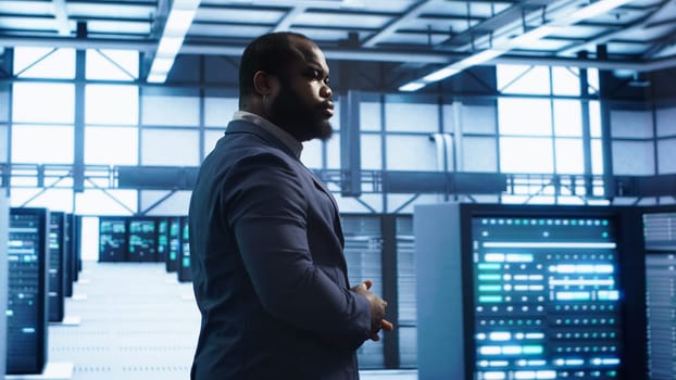 African american technician walking in data center, monitoring systems to identify errors. Man in server hub location doing diagnosis, looking for infrastructure bugs in need of repairing, camera A