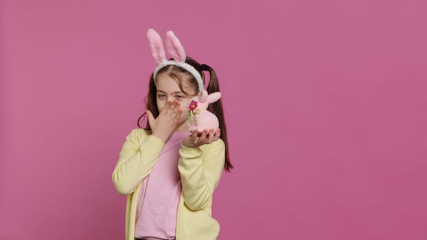 Jolly schoolgirl holding a cute rabbit toy and blowing kisses in studio, being adorable with festive decorations for easter holiday event. Happy child wearing bunny ears and pigtails. Camera A.