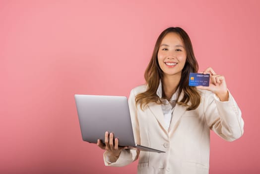 Portrait of Asian beautiful young woman customer showing credit card for online payment shopping transactions cashback loan on laptop studio shot isolated on pink background