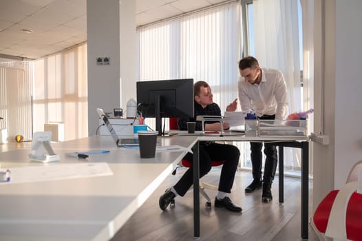 Caucasian man standing at colleague's desk discussing work