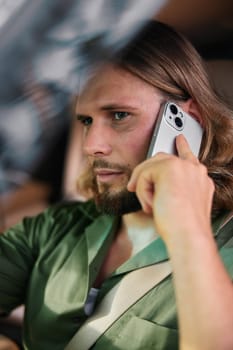 A man with long hair is engaged in a phone conversation while driving, wearing a green shirt, radiating concentration