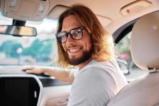 Happy young man with long hair and glasses in a car, looking back with a smile, creating a warm atmosphere