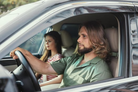 A man and woman sit in a car, the man driving, focused on the road ahead, while the woman gazes away, lost in thought