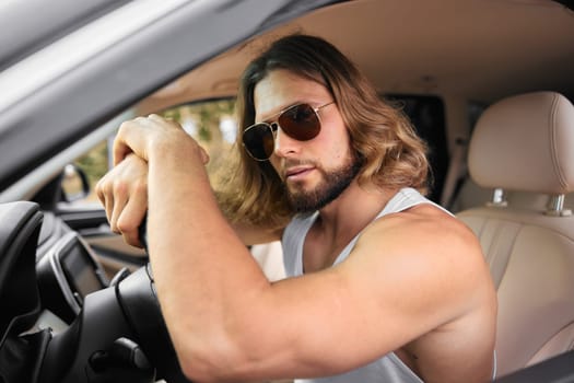 A stylish male with long hair and sunglasses, sitting in the driver s seat of a car, exuding confidence and sophistication against a neutral interior backdrop