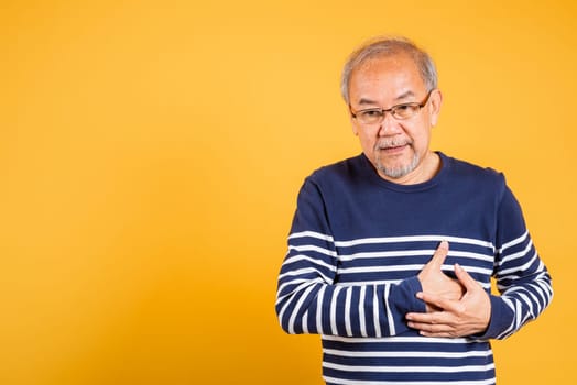 Portrait Asian old man with glasses holds hands on chest studio shot isolated yellow background, senior man pensioner with grey hair heart disease suffering from chest pain