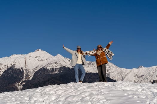 Two women joyfully pose on a snow-covered mountain peak against a backdrop of a clear blue sky and majestic snow-capped mountains.