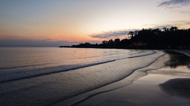 Small waves gently washing a tropical beach at sunset with orange, pink and purple colors in the sky and dark silhouettes of trees and buildings in the background