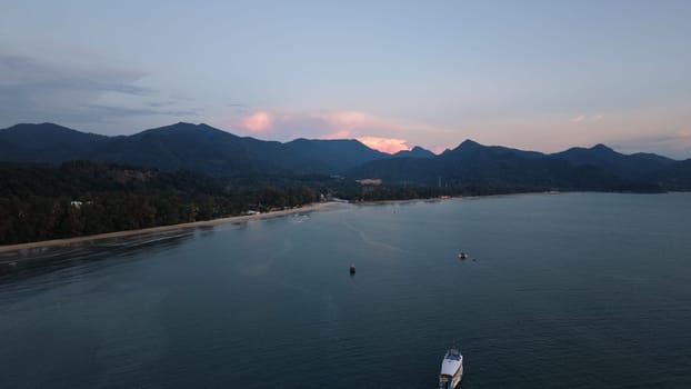 Stunning aerial view capturing boats floating on calm water near a tropical beach at sunset, framed by lush vegetation and majestic mountains
