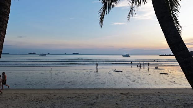 Tourists are walking and playing on a beautiful tropical beach at sunset, with palm trees framing the scene and a yacht sailing in the distance