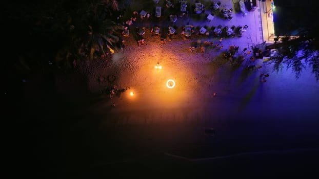 Aerial view capturing a vibrant beach resort at night, with a mesmerizing fire show lighting up the sandy shore as tourists gather along the coastline, enjoying the festive ambiance