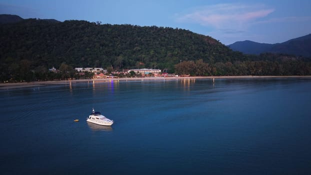 Motor yacht floating near a tropical beach at dusk in thailand, with lights reflecting on the calm water and forested hills in the background